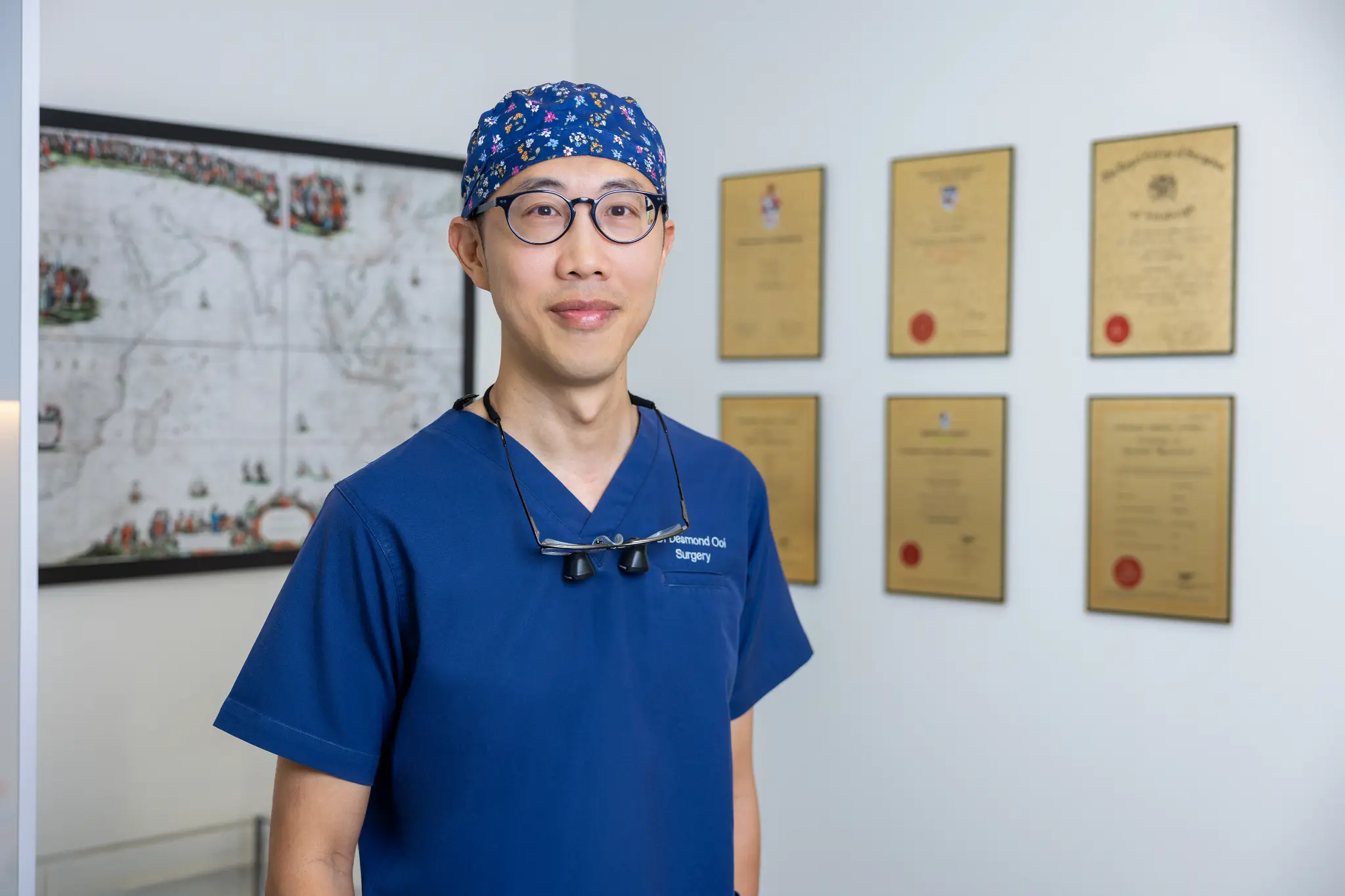Dr Desmond Ooi in surgical scrubs with loupes, standing before his framed fellowship certificates